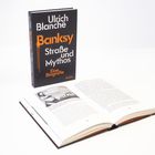 "Ulrich Blanché, Banksy: Straße und Mythos, Eine Biografie, Reclam." Ein aufgestelltes Buch mit einem geöffneten Exemplar davor.