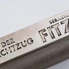 Text: "Der Nachtzug", "Sebastian Fitzek". Geprägter Schriftzug auf einer metallicfarbenen Oberfläche.