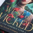 "West of Wicked: Great and Terrible Land I," mit einer Frau in einem blauen Kleid und roten Blüten im Vordergrund.