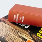 Ein orangener Buchrücken mit der Aufschrift "Colson Whitehead Cool Machine" liegt auf einem Buch mit Gebäudeabbildung.