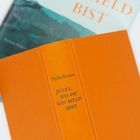 Buchtitel: "Jetzt, wo du ein Held bist" von Thilo Krause. Ein orangefarbenes Buch liegt auf einem anderen Buch.