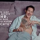 Schauspieler Brix Schaumburg liegt lächelnd auf einem lilafarbenen Kissen mit einem leeren Glas in der Hand.
