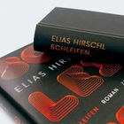 Text: "Elias Hirschl, Schleifen, Roman." Zwei Bücher, eines liegt oben, Gestaltung in Schwarz mit orange-rotem Muster.