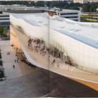 Innenhof einer modernen Bibliothek, geschwungene Architektur, Menschen sitzen auf der Terrasse, Vegetation sichtbar.