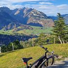 Ein Fahrrad lehnt an einer Holzbank vor einer malerischen Berglandschaft, umgeben von grünen Wiesen und Tannen.