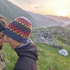 Zwei Personen stehen mit Stirn an Stirn in einer grünen Berglandschaft bei Sonnenuntergang, im Hintergrund ein Zelt.