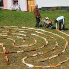 Kein Text im Bild. Zwei Personen legen Holzscheite in einem spiralförmigen Muster auf Gras. Eine Person schiebt eine Schubkarre.