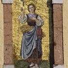 Eine Heilige mit Heiligenschein hält eine Sichel und Ähren, dargestellt in einem goldenen Mosaik.
