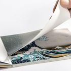 Eine Hand blättert durch ein kleines Buch mit einer Illustration einer großen Welle und einem Berg im Hintergrund.