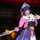 Eine Person in detailreichem, buntem Cosplay trägt ein violettes Gewand mit Verzierung und hält eine dekorative Waffe.