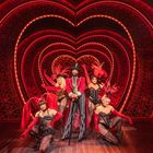 Fünf Personen in auffälligen roten Kostümen stehen vor einem leuchtenden, herzförmigen Bühnenbild.