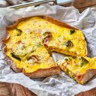 Ein Stück Quiche mit gelber Füllung, Brokkoli und Schinken liegt auf Backpapier. Ein blau-weißer Teller im Hintergrund.