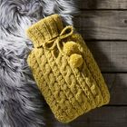 Ein gelber Strick-Wärmflaschenbezug mit Zopfmuster und Pompons liegt auf einem grauen, flauschigen Fell auf Holzdielen.
