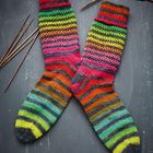 Zwei bunte gestrickte Socken mit Streifenmuster in Regenbogenfarben, umgeben von Stricknadeln auf dunklem Hintergrund.