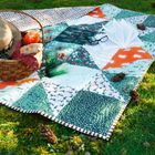 Auf einer bunten Decke im Gras liegt ein offenes Buch mit Brille. Daneben ein Picknickkorb mit Brot und Getränken.