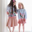Zwei Mädchen stehen lachend nebeneinander, tragen ähnliche gestrickte Pullover mit Blumenmotiven und rosa Röcken.