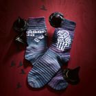 Zwei gestrickte Socken mit Mustern, darunter eine Eule. Kleine Hexenhüte und -silhouetten umrahmen sie.
