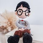 Ein handgemachtes Stoffpüppchen mit Brille und schwarzem Haar sitzt auf einem weißen Tisch, im Hintergrund Pampasgras.