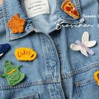 Season 2: Broschen & Pins. Jeansjacke mit bunten, gehäkelten Patches, darunter Pizza und ein Blatt.
