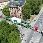 Große Ladung auf einem langen Transportfahrzeug mit „Kübler“ Beschriftung auf Straßenkreuzung, umgeben von Bäumen und Gebäuden.