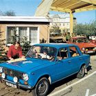 Nummernschild: 18G-U-94. Ein Mann in rotem Hemd liest eine Karte auf einer blauen Limousine vor einer Tankstelle.
