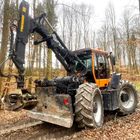 Ein Traktor mit großem Greifarm fährt durch einen herbstlichen Wald mit kahlen Bäumen.