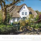 "1851" auf weißem Haus. Reetdach, Garten, Bäume, großes rostiges Ankerobjekt seitlich, Monatskalender unten: März.