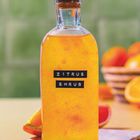 Text: "ZITRUS SHRUB". Klare Flasche mit Korken, gefüllt mit gelber Flüssigkeit. Hintergrund verschwommen, mit Orangen.