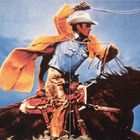 Ein Cowboy in gelber Jacke und Hut schwingt ein Lasso auf einem galoppierenden Pferd vor blauem Himmel.