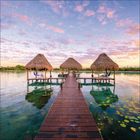 Ein Steg führt zu drei Strohhütten über einem ruhigen, klaren Wasser mit Seerosen, unter einem Himmel mit rosa Wolken.