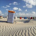 Strandkörbe auf hellem Sand, Nummern wie 58, 29, und 25 erkennbar, unter blauem Himmel mit weißen Wolken.