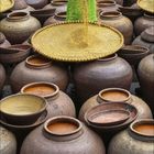 3, 2023. Zahlreiche große, braune Tongefäße. Person in grünem Gewand und Hut verteilt Getreide.