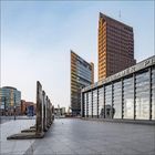 Text: "BAHNHOF POTSDAMER PLATZ". Moderne Gebäude mit Glasfassaden, Reste der Berliner Mauer im Vordergrund.