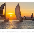 3. März. Segelboote auf Wasser bei Sonnenuntergang, kräftige Farben, ruhige Stimmung, Berge im Hintergrund.