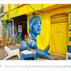 "La Bohème" in Blau, großes Wandgemälde mit Porträt in Blau und Weiß, gelbe Fassade, blaue und weiße Stühle davor.