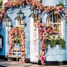 CHEYNE. Dekorierte Hausfassade mit blauen Wänden und Blumenarrangements um Fenster und Türen, Kalenderblatt März.