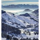 02 JANUAR. Schneebedeckte Berge, bewaldete Hänge, nebelige Täler, im Hintergrund eine beeindruckende Gebirgskette.