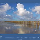 März 2027, Foto eines sandigen Strands mit Holzpfählen im Wasser unter blauem Himmel und Wolken.