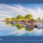 März 2027. Segelboote liegen in einem ruhigen, sonnigen Hafen, umgeben von blühenden Bäumen und Gebäuden am Ufer.