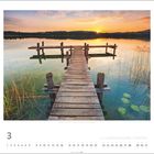 März 2022. Kalenderbild mit Holzsteg, Sonnenuntergang über einem ruhigen See, Lilienblättern und Schilf.