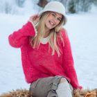 Eine lächelnde Frau in rotem Pullover und weißem Hut sitzt im Schnee. Im Hintergrund schneebedeckte Bäume.
