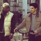 Zwei junge Männer gehen draußen, einer schiebt ein Fahrrad. Beide tragen Jacken und unterhalten sich.