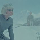 Eine Frau in grau kniet im Schnee, trägt einen glitzernden Kopfschmuck. Im Hintergrund ist eine verschwommene Gestalt erkennbar.