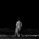 Keine sichtbaren Texte. Eine Person in gemustertem Gewand steht am nächtlichen Strand, blickt aufs Meer.