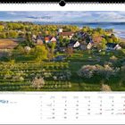 Märzkalender mit Landschaftsbild: Dorf in Frühlingslandschaft, Blütenbäume, Hintergrund Schweizer Alpen.