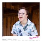 Text: Melvin, September. Junge mit Brille und Hawaiihemd lächelt herzlich, Holzstruktur im Hintergrund.