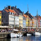 Bunte Häuserreihe am Wasser, Segelboote im Hafen, Menschen an der Promenade, blauer Himmel.