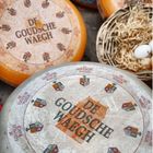 „De Goudsche Waeght“ steht auf der Oberseite von Käselaiben. Daneben liegt ein Korb mit Stroh und Eiern.