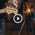 „Guitarschool.at“ unten, ein Smartphone zeigt eine E-Tuner-App. Eine Person stimmt eine E-Gitarre.