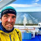 „honi mara“ auf einem Stirnband. Ein Mann auf einem Schiff mit blauem Meer im Hintergrund. Er trägt eine gelbe Jacke.
